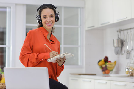 Happy latin woman learn cooking food while checking recipe from internet with computer in kitchen. Young female with headphone prepare delicious meal and listen music. Healthy and lifestyle conceptの写真素材