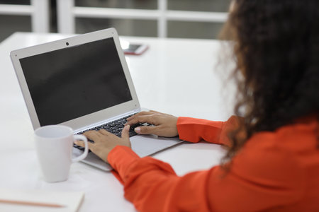 Rear view smart and active latin woman hands sitting and using internet on computer while using smart mobile phone with a cup of coffee in kitchen. Anywhere and work. Lifestyle with technologyの写真素材