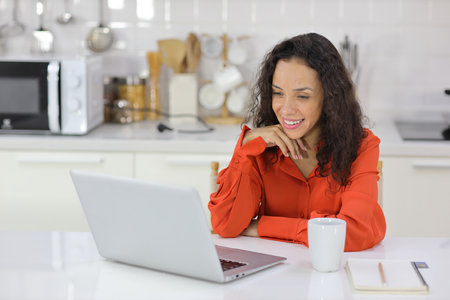 Smart and active latin woman sitting and using internet on computer while using smart mobile phone with a cup of coffee in kitchen. Anywhere and work. Lifestyle with technologyの写真素材