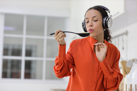 Happy latin woman cooking, smelling and tasting food in kitchen. Beautiful young female with headphone preparing delicious meal while listen music at home. Healthy and lifestyle conceptの写真素材