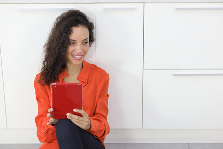 Happy latin woman sitting and using internet on tablet in kitchen room. Businesswoman looking at camera with smiling. Lifestyle with technologyの写真素材