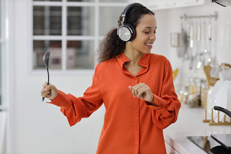 Happy latin woman cooking, smelling and tasting food while dancing in kitchen. Beautiful young female with headphone preparing delicious meal while listen music at home. Healthy and lifestyle conceptの写真素材
