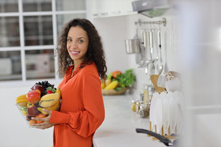 Happy latin woman standing and holding fruits basket while smiling and looking at camera in kitchen. Young female preparing delicious meal at home. Healthy and lifestyle conceptの写真素材