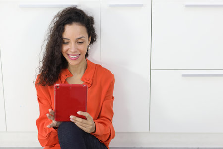 Happy latin woman sitting and using internet on tablet in kitchen room. Businesswoman looking at camera with smiling. Lifestyle with technologyの写真素材