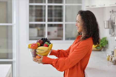 Happy latin woman standing and holding fruits basket while smiling and looking away in kitchen. Young female preparing delicious meal at home. Healthy and lifestyle conceptの写真素材