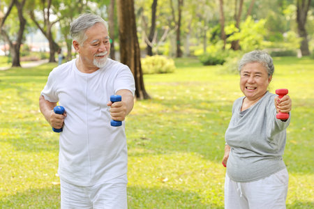 Happy and smiling asian senior couple doing arm work out and lifting dumbbell exercise with relaxation for healthy in park outdoor after retirement. Health care elderly outdoor lifestyle concept.の写真素材