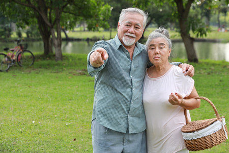 Happy asian senior man and woman walking and hugging while pointing something with picnic basket in garden outdoor. Lover couple going to picnic at the park. Happiness marriage lifestyle concept.の写真素材