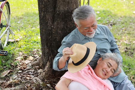 Portrait of happy asian senior man and woman lying on lap and embracing with bicycle and hat in summer garden outdoor. Lover couple going to picnic at the park. Happiness marriage lifestyle concept.の写真素材