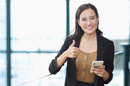 Portrait of confident young asian businesswoman researching and planning work with happy smiling face while showing thum up at business meeting in modern office building and city backgroundの写真素材
