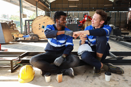Happy teamwork technician engineer in safety workwear make fist bumping celebrate successful together completed deal commitment during eating with sandwich and coffee at industry manufacturing factoryの写真素材