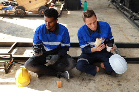 Happy teamwork technician engineer in safety workwear having lunch and eating sandwich and coffee at industry manufacturing factoryの写真素材