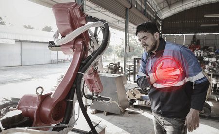 Young technician engineer or worker man in safety uniform suffering from chest pain or heart attack from working accident at industrial robotic factory. First aid, healthy or insurance at work conceptの写真素材