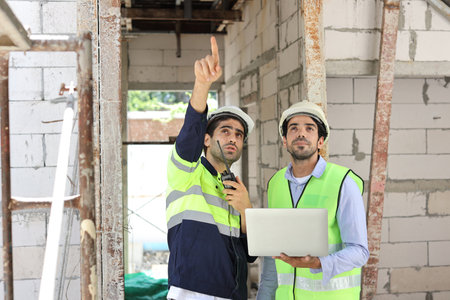 Two technician civil engineer or specialist inspector pointing, discussing and plan work with laptop, blueprint and walkie talkie radio together with at Industrial building site. Construction conceptの写真素材