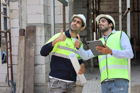 Two technician civil engineer showing thumb up while discussing, brainstorm and planning work with laptop, blueprint and walkie talkie radio together at Industrial building site. Construction conceptの写真素材
