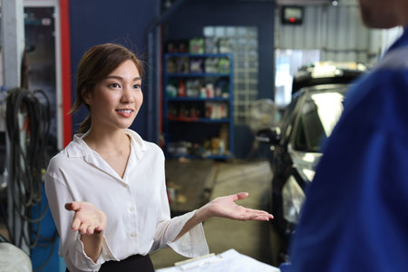 Young woman customer listening her car repairs problemsÂ condition from auto mechanic. Man technician car mechanical in uniform show car maintenance service report on clipboard at repair garage stationの写真素材