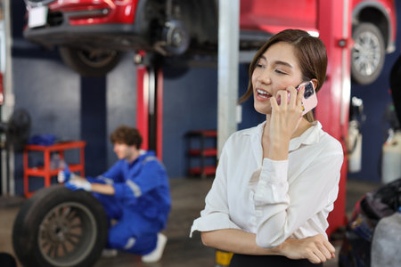 Young asian woman customer using smart mobile phone and calling insurance agent to get help or support at repair garage station with mechanic in uniform working blurred backgroundの写真素材