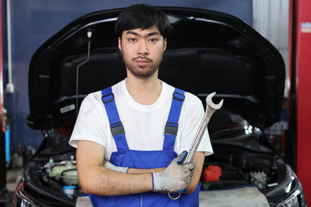 Asian technician engineer or worker man uniform standing and holding wrench checking maintenance a car service with confident at repair garage station. Concept of car center repair service.の写真素材