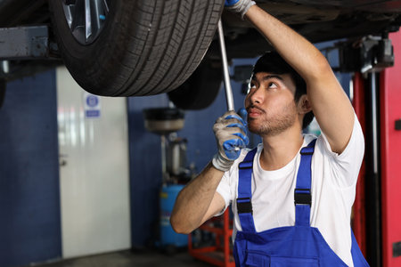 Man technician car mechanic in uniform check or maintenance a lifted car service at repair garage station. Worker holding wrench and fixing vehicle with flashlight. Car center repair service conceptの写真素材