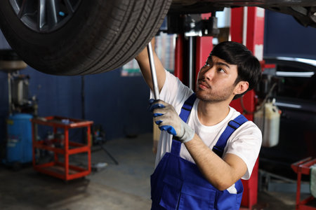 Man technician car mechanic in uniform check or maintenance a lifted car service at repair garage station. Worker holding wrench and fixing vehicle with flashlight. Car center repair service conceptの写真素材