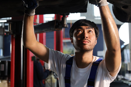 Man technician car mechanic in uniform check or maintenance a lifted car service at repair garage station. Worker holding wrench and fixing vehicle with flashlight. Car center repair service conceptの写真素材