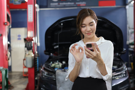 Young woman customer using smart mobile phone and calling insurance agent to get help or support at repair garage station with breakdown car blurred background. Concept of car center repair service.の写真素材