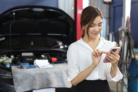 Young woman customer using smart mobile phone and calling insurance agent to get help or support at repair garage station with breakdown car blurred background. Concept of car center repair service.の写真素材
