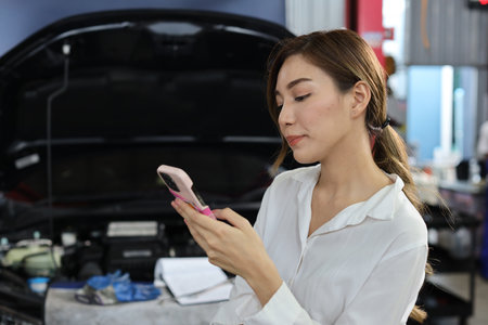 Young woman customer using smart mobile phone and calling insurance agent to get help or support at repair garage station with breakdown car blurred background. Concept of car center repair service.の写真素材