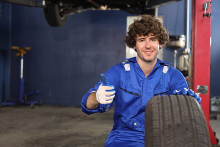 Man technician car mechanic in uniform holding tire for replacement or changeing at repair garage. Happy caucasian worker showing thumb up and looking at camera. Concept of car center repair service.の写真素材