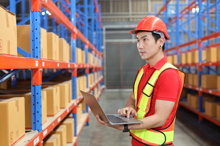 Warehouse workers man with hardhats and reflective jackets using computer, walkie talkie radio and cardboard while controlling stock and inventory in retail warehouse logistics, distribution centerの写真素材