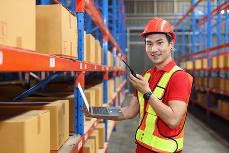 Warehouse workers man with hardhats and reflective jackets using computer, walkie talkie radio and cardboard while controlling stock and inventory in retail warehouse logistics, distribution centerの写真素材
