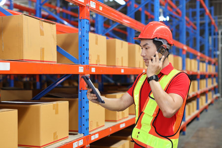 Warehouse workers man with hardhats and reflective jackets using computer, smart mobile phone and tablet while controlling stock and inventory in retail warehouse logistics, distribution centerの写真素材