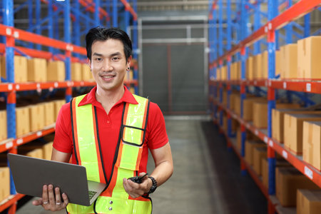 Warehouse workers man with hardhats and reflective jackets using computer, walkie talkie radio and cardboard while controlling stock and inventory in retail warehouse logistics, distribution centerの写真素材