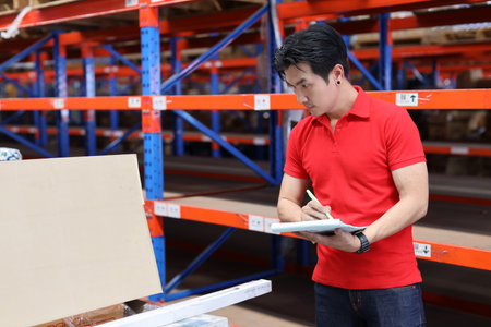 Warehouse workers young asian man working and using tablet, walkie talkie radio and cardboard while controlling stock and inventory in retail warehouse logistics, distribution centerの写真素材
