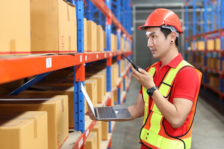 Warehouse workers man with hardhats and reflective jackets using computer, walkie talkie radio and cardboard while controlling stock and inventory in retail warehouse logistics, distribution centerの写真素材