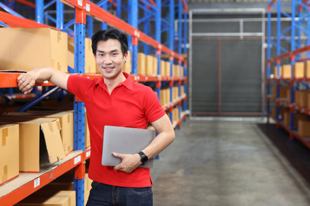 Portrait of warehouse workers young asian man standing and using computer while looking at camera and controlling stock and inventory in retail warehouse logistics, distribution centerの写真素材