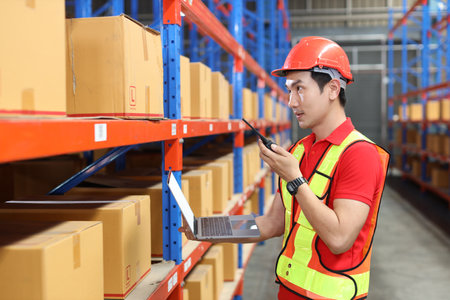 Warehouse workers man with hardhats and reflective jackets using computer, walkie talkie radio and cardboard while controlling stock and inventory in retail warehouse logistics, distribution centerの写真素材