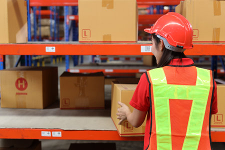 Rear view warehouse workers woman with hardhats and reflective jackets carrying a large box for delivery to production stock and inventory in retail warehouse logistics, distribution centerの写真素材