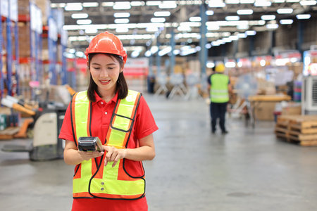 Warehouse workers woman with hardhats and reflective jackets while holding scanner barcode and looking at camera with production stock and retail warehouse logistics distribution center backgroundの写真素材