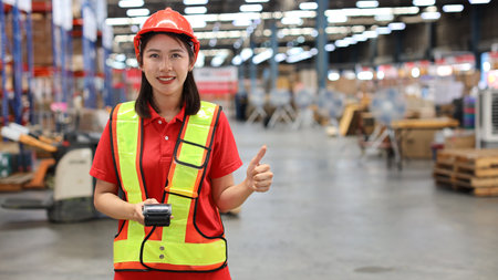 Warehouse workers woman with hardhats holding scanner barcode while showing thumb up and looking at camera with production stock and retail warehouse logistics distribution center backgroundの写真素材