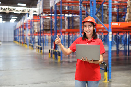 Portrait of warehouse workers young asian woman standing and using computer while showing thumb up and controlling stock and inventory in retail warehouse logistics, distribution centerの写真素材