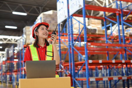 Warehouse workers woman with hardhats and reflective jackets using computer, walkie talkie radio and cardboard while controlling stock and inventory in retail warehouse logistics, distribution centerの写真素材