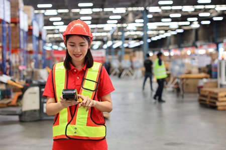Warehouse workers woman with hardhats and reflective jackets while holding scanner barcode and looking at camera with production stock and retail warehouse logistics distribution center backgroundの写真素材