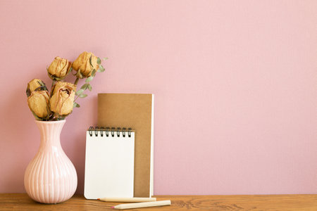 Empty notebook with vase of dry rose flowers on wooden table. pink backgroundの写真素材