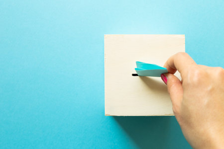 Hand putting voting paper in Ballot box on blue background. top view, copy space