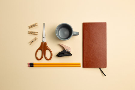 Notebook, mug cup, pencil, stapler, clip, scissors on beige background. flat lay, top view, copy space. Work and study placeの写真素材