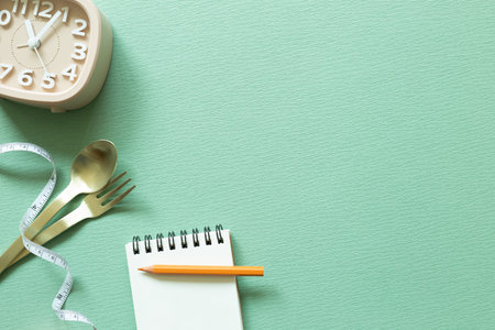 Fork and spoon with measuring tape, note pad, clock on green background. Diet concept