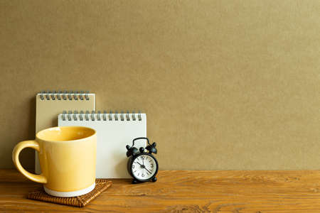 Notebook, mug cup, clock on wooden desk. brown wall background. workspaceの写真素材