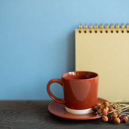Brown cup of coffee, notebook, dry flowers on wooden desk. blue wall background. workspaceの写真素材