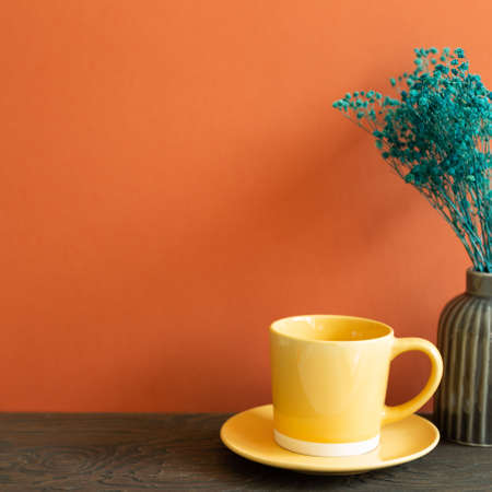 Yellow coffee cup and vase of green dry flowers on wooden table. red wall backgroundの写真素材
