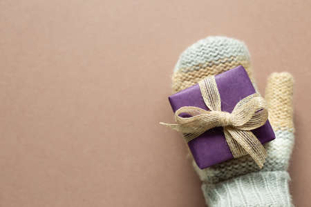 Hand in winter knitted gloves with purple gift box on brown background. anniversary, christmas, greeting concept. top view, copy spaceの写真素材